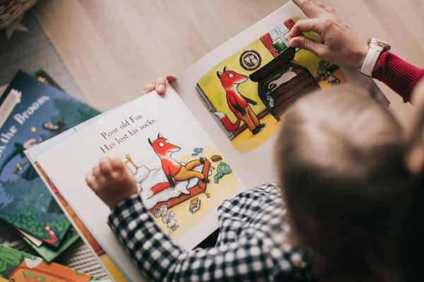 A child sits with an adult looking at an illustrated storybook about a fox who has lost his socks, with colorful pages and other children’s books scattered nearby.
