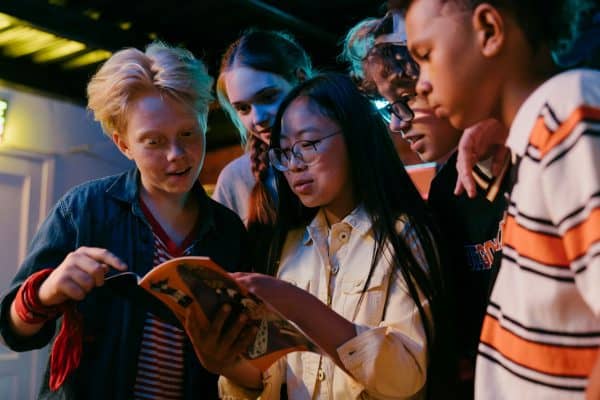 A group of kids stand closely together reading a book, looking engaged and curious under colorful indoor lighting.