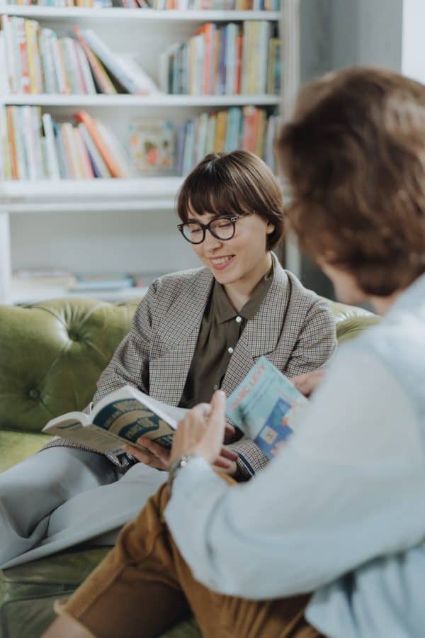 Two people sit on a green couch in a library reading books together, with shelves of colorful books in the background; one person smiles while holding an open book.